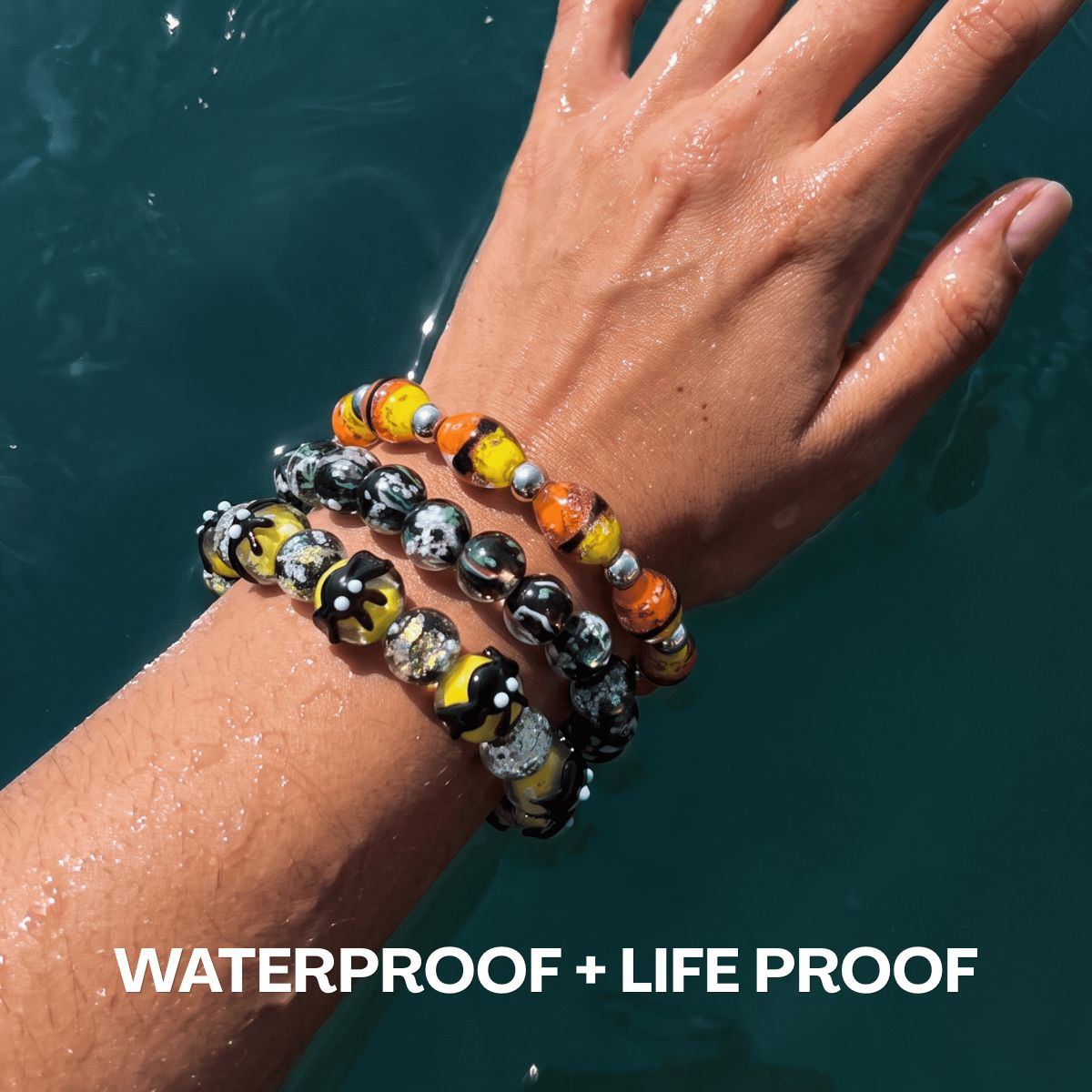 Stacked waterproof beaded bracelets in yellow, orange, black and green glass on a wet wrist in blue water.