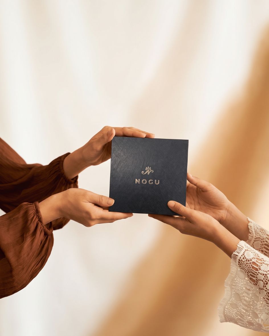 Hands exchange a navy blue NOGU jewelry gift box with a gold logo against a warm, softly lit background.