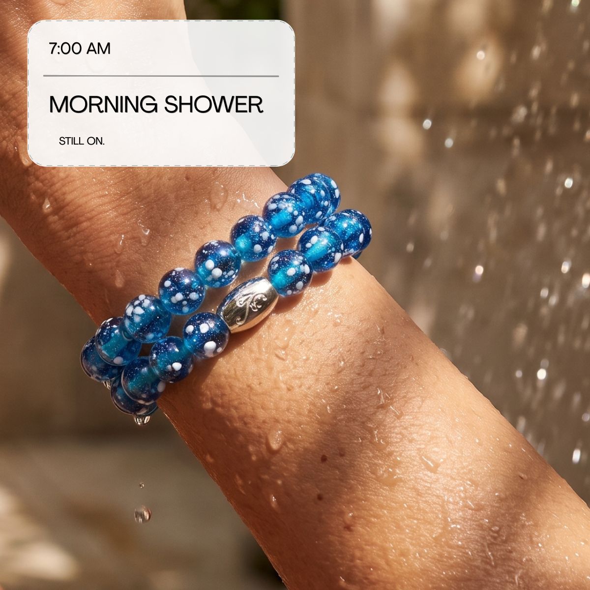 A close-up of a person's wrist wearing two blue beaded bracelets with white speckles and a silver accent bead, photographed outdoors with water droplets and a blurred warm-toned background. A white text box in the upper left reads '7:00 AM MORNING SHOWER STILL ON'.