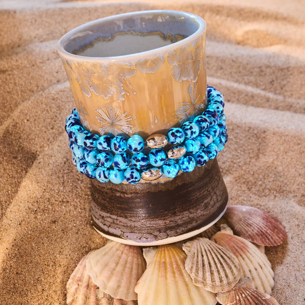 Blue Seaspray Firefly Glass Bracelet with blue hand-blown beads and sterling silver accents displayed on ceramic cup on sand by NOGU