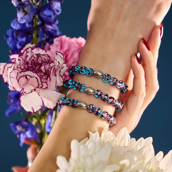 Purple Elixir bracelets with glowing purple and blue Firefly Glass beads and silver accents stacked on wrist with flowers by NOGU